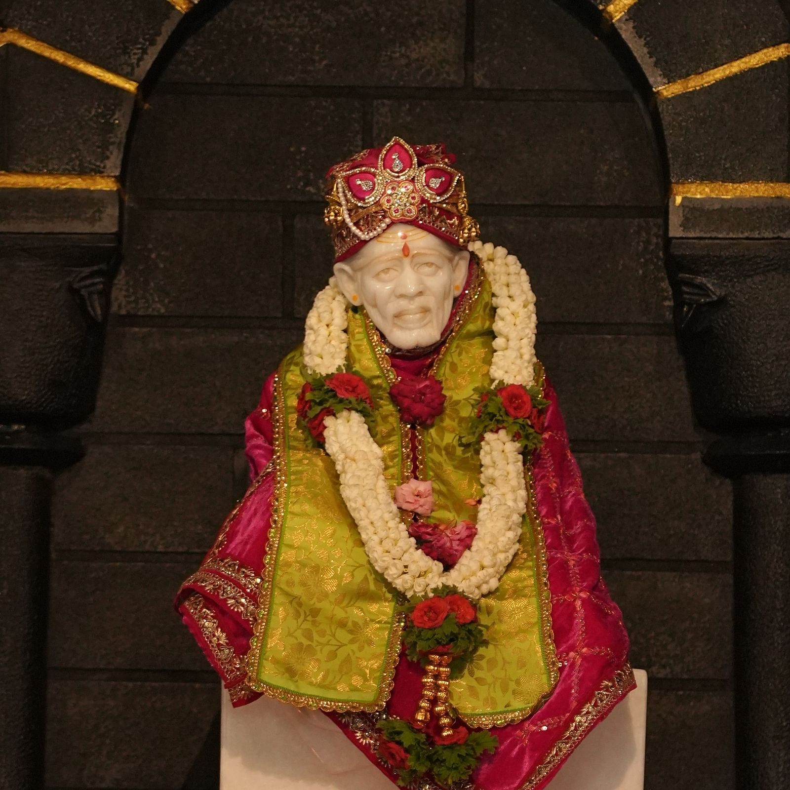 Statue of Shirdi Sai Baba adorned with garlands, royal attire, and lit oil lamps. | Hoysala Village Resort